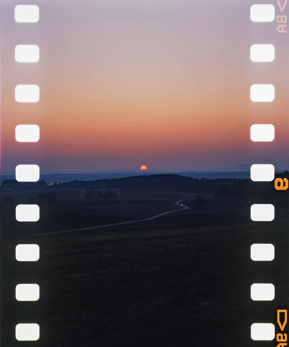 The sun sets over a rural landscape with a winding road and distant hills.