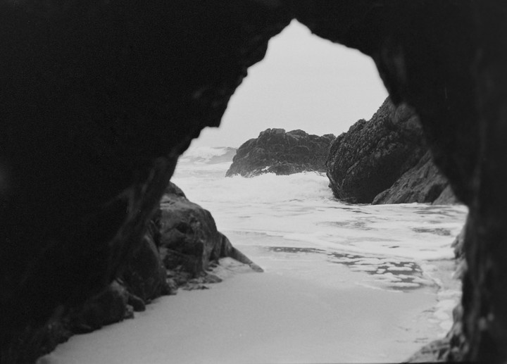 Waves crash against rocks on a sandy beach, viewed through a dark rock archway.