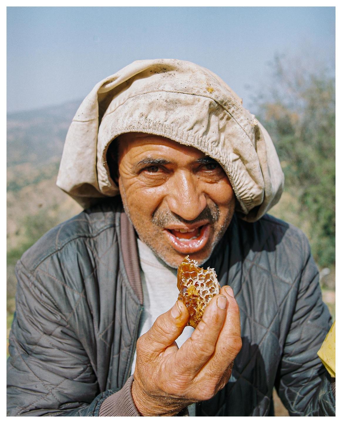 A man wearing a light-colored cap holds a piece of honeycomb to his mouth.