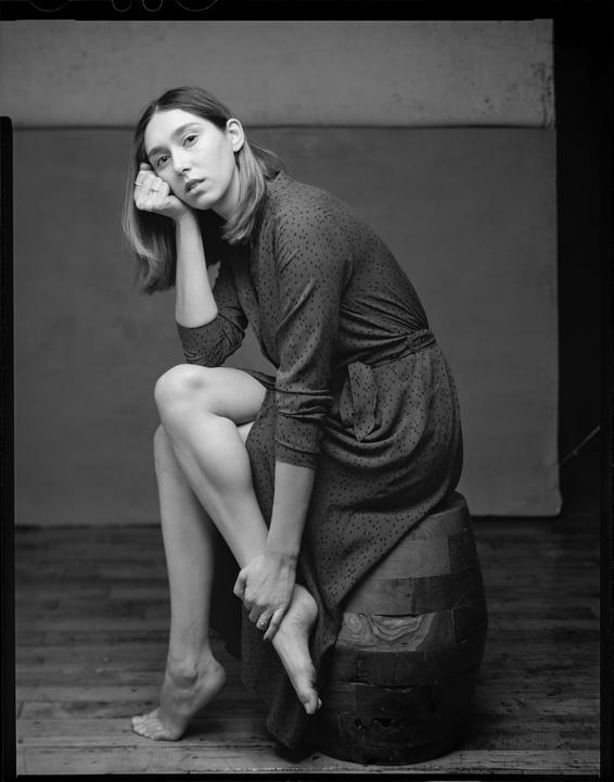 A woman in a patterned dress sits barefoot on a wooden stool, resting her chin on her hand.