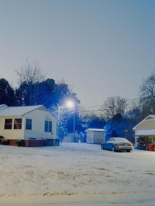 A suburban street is covered in snow with houses and a car visible under a bright street light.