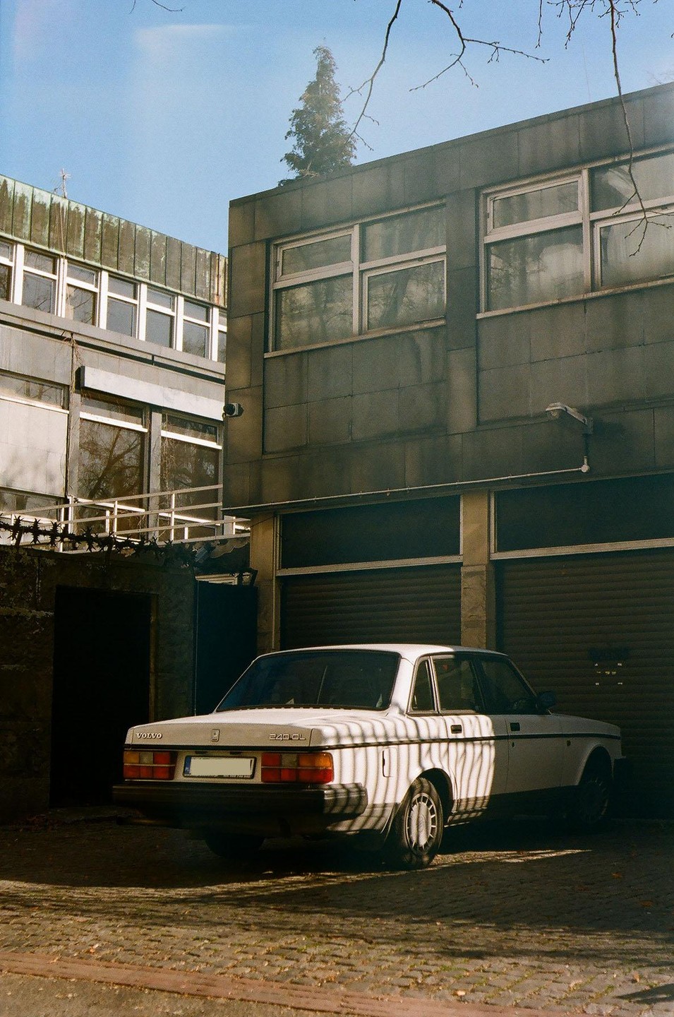 A white Volvo 240 GL is parked on a cobblestone street next to a building with closed garage doors, with shadows cast across the car.