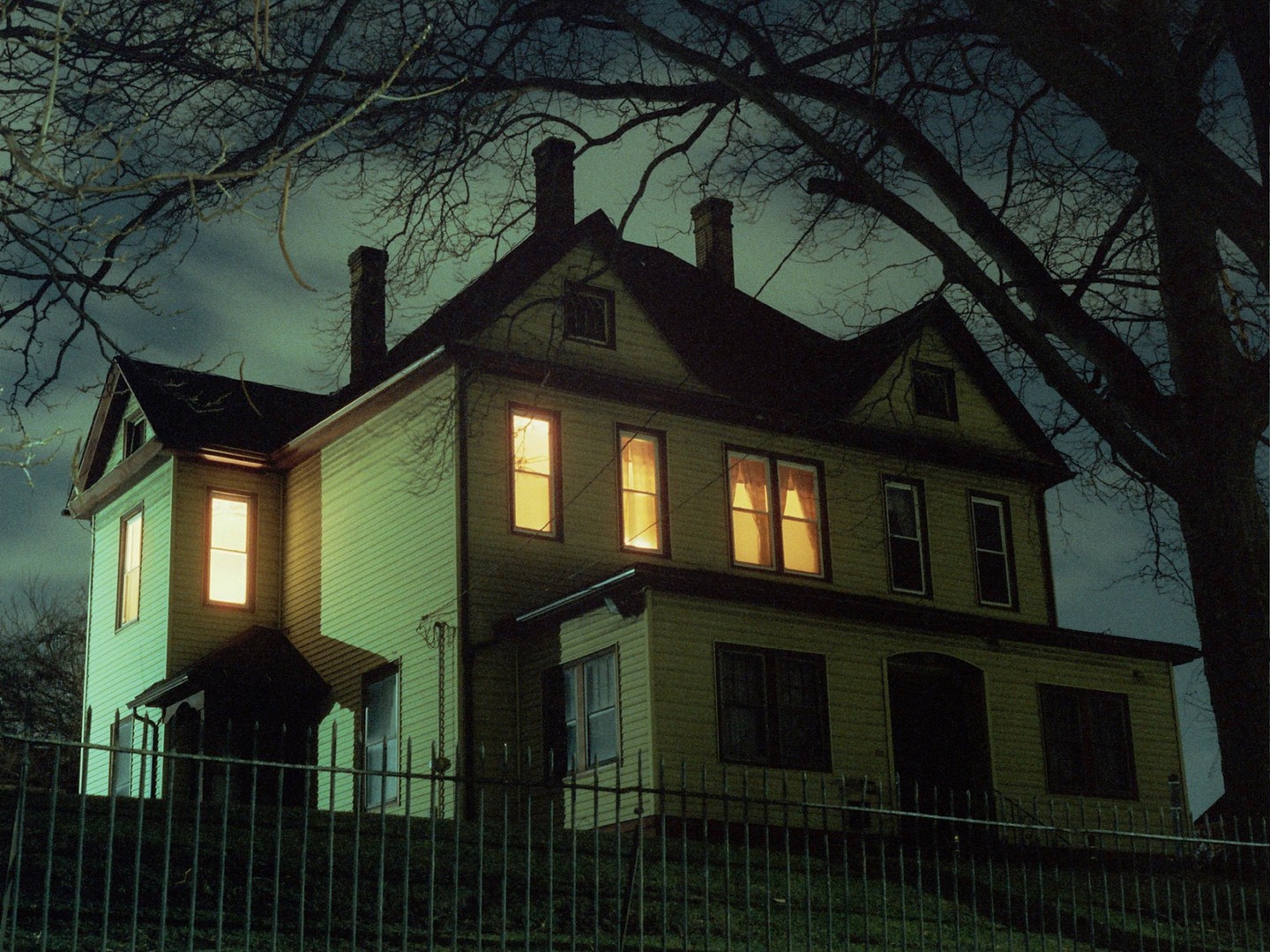 A large house with illuminated windows stands behind a fence at night, framed by bare tree branches.