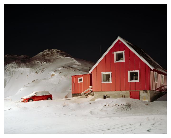 A red house and a red car are surrounded by snow at night with mountains in the background.