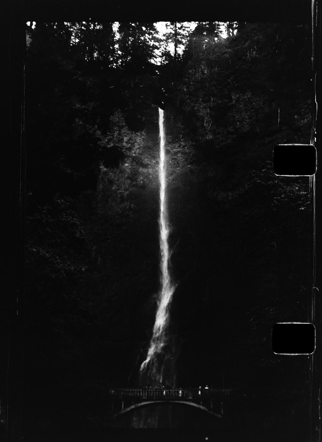 A tall waterfall cascades down a dark, rocky cliff face, with a bridge and people visible at its base.