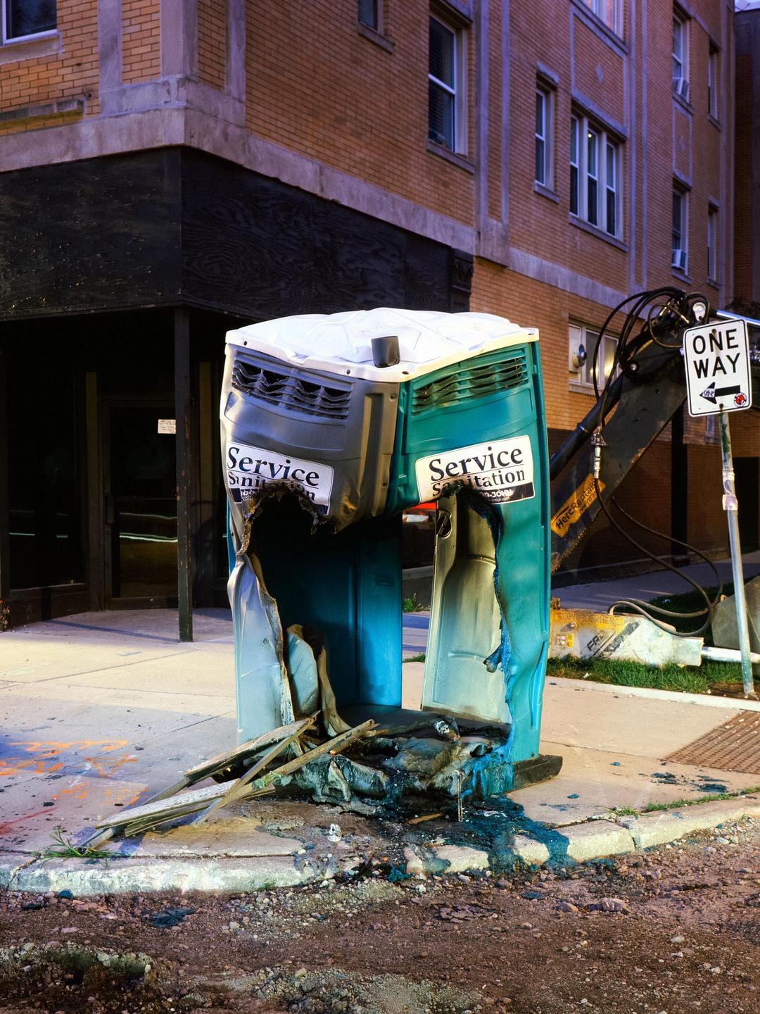 A destroyed portable toilet sits on a sidewalk next to a brick building at night.