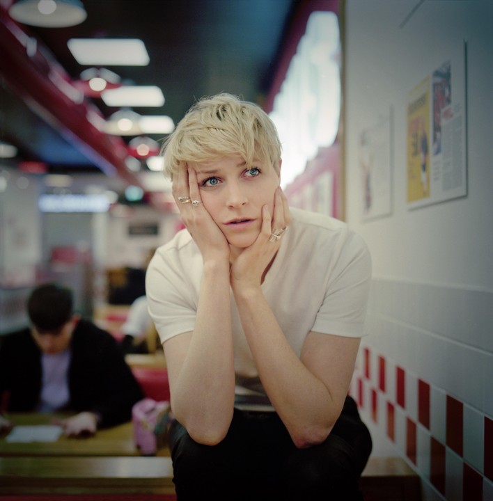 A woman with short blonde hair and blue eyes rests her face in her hands while sitting in a diner.