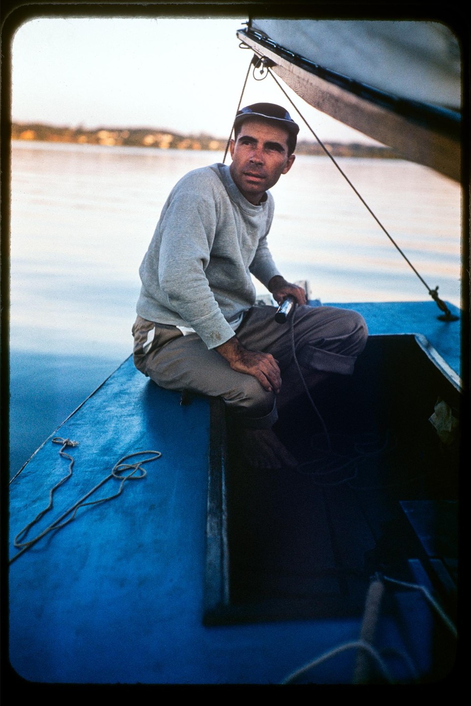 A man wearing a cap and sweatshirt sits on the edge of a blue boat, looking towards the right with water and land in the background.