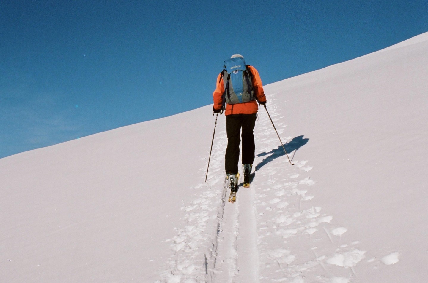A person wearing an orange jacket and a blue backpack skis up a snow-covered mountain under a clear blue sky.