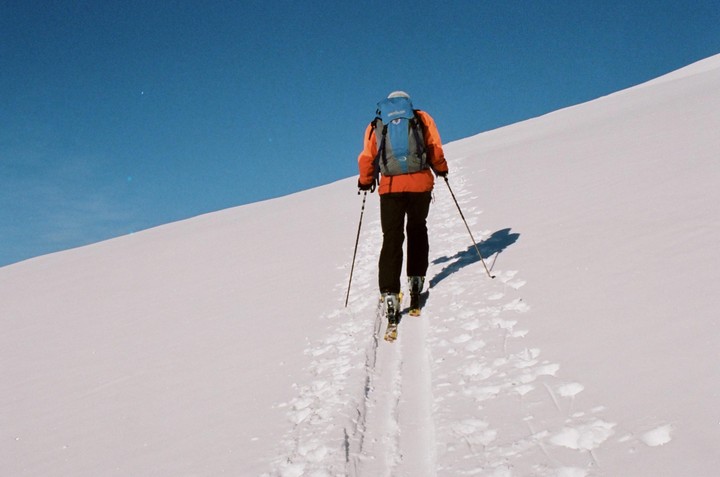 A person wearing an orange jacket and a blue backpack skis up a snow-covered mountain under a clear blue sky.