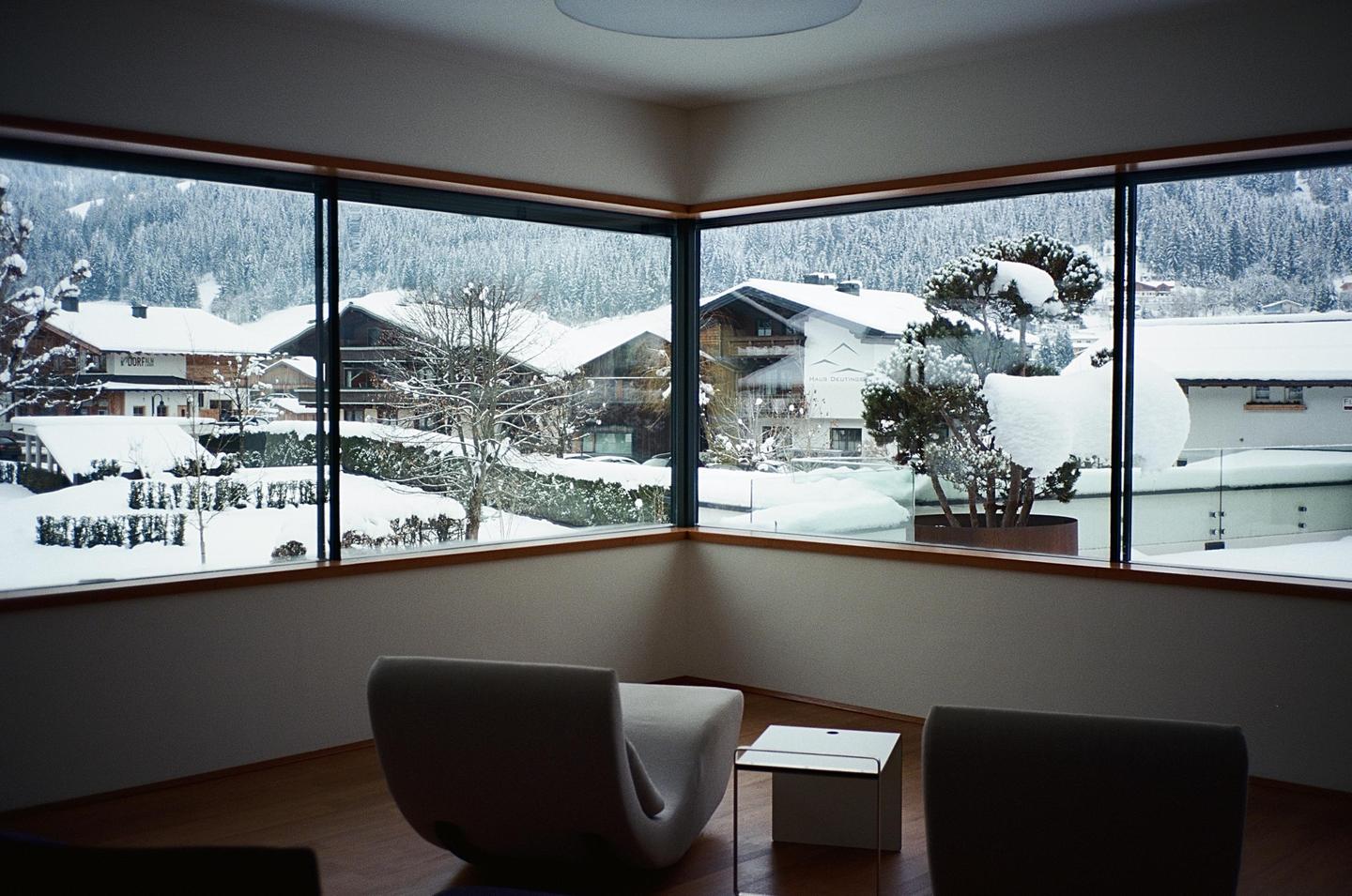 A modern room with two chairs and a small table looks out through large windows onto a snow-covered village and forest.