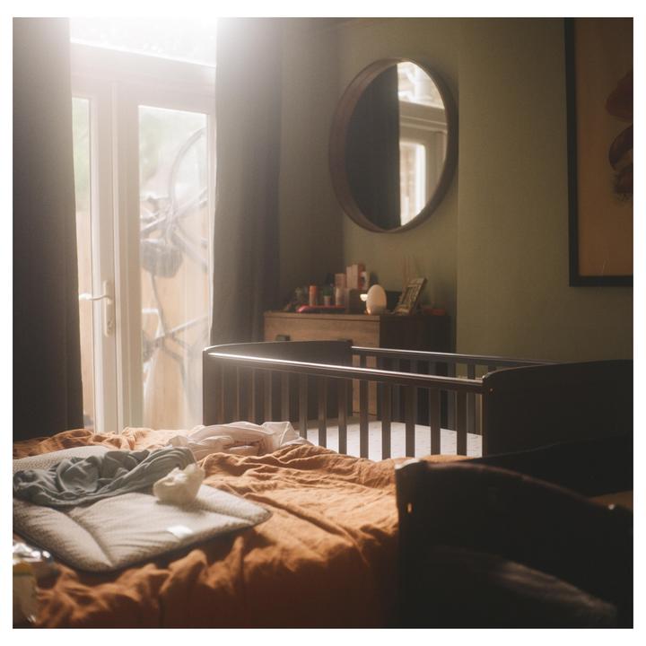 A baby's crib stands in a sunlit room with a bed in the foreground.