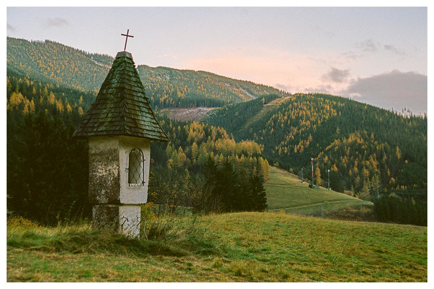 A small shrine with a cross on its roof stands in a grassy field with a forest and mountains in the background.
