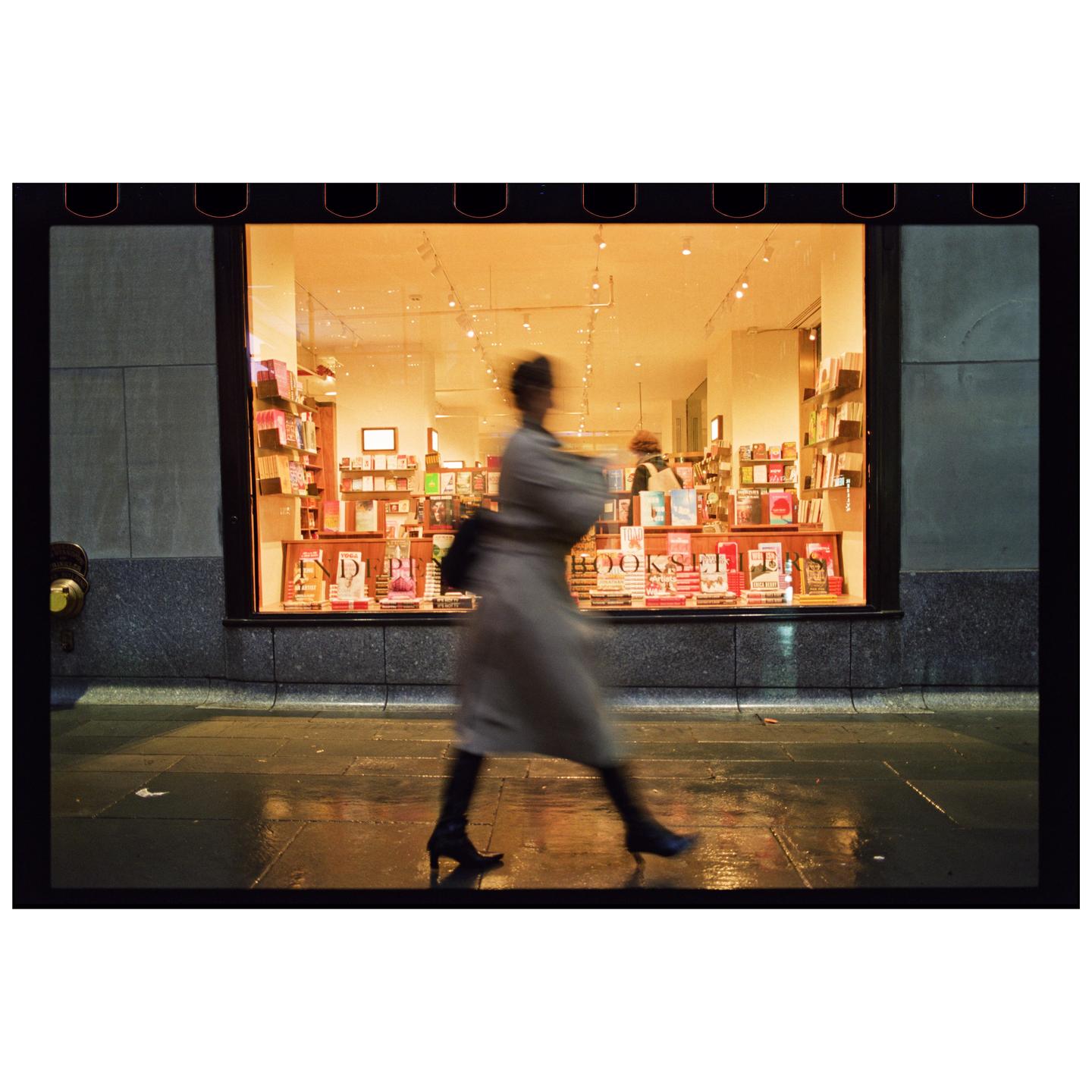 A blurry woman walks past a brightly lit bookstore window at night.