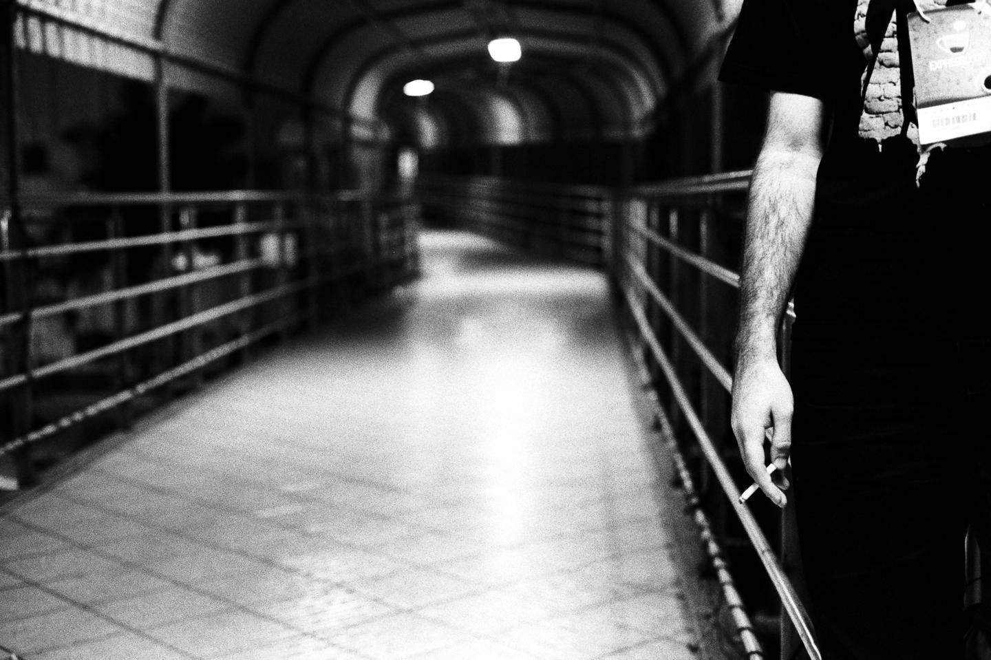 A man holds a cigarette while standing in a long, dimly lit tunnel with railings on both sides.