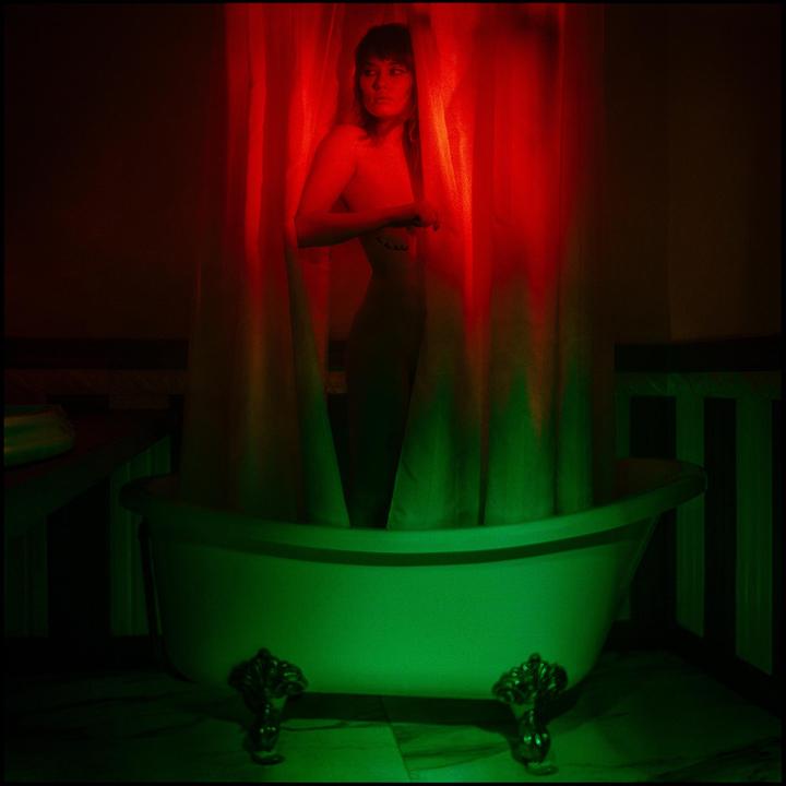 A woman stands behind a shower curtain in a clawfoot bathtub, illuminated by red and green light.