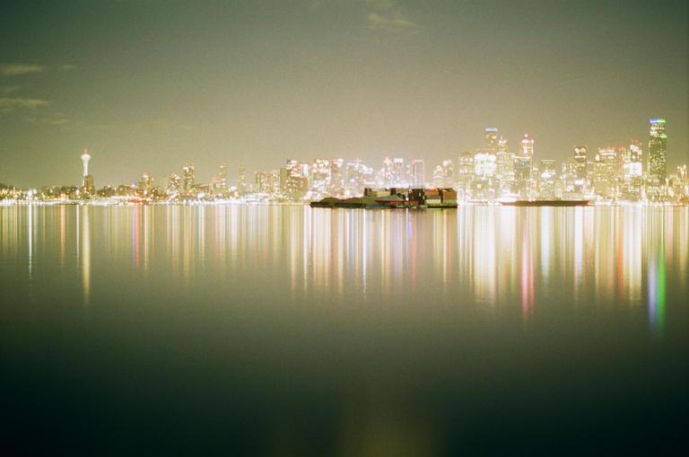 A city skyline with a prominent tower is reflected in the calm water at night, with a barge visible in the middle ground.