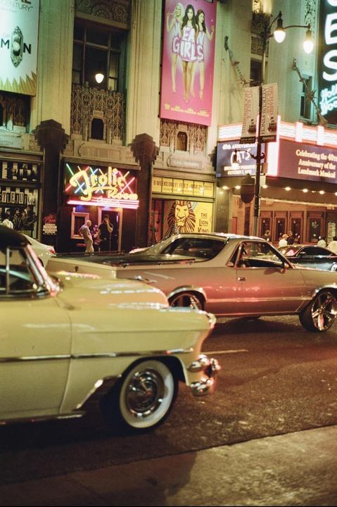 Two classic cars are parked on a street at night, with neon signs and theater posters visible in the background.