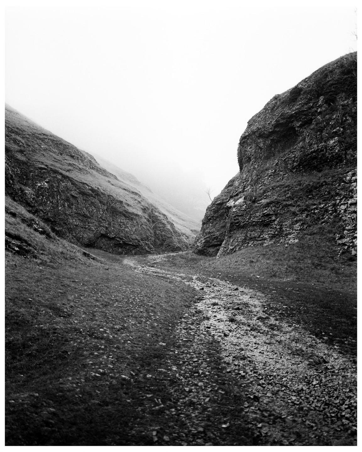 A rocky path winds through a valley between two steep, grassy hills under a hazy sky.