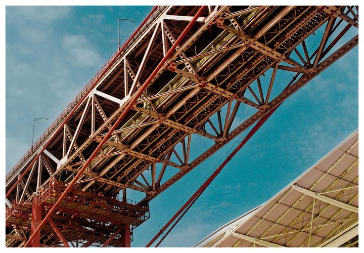 A large red and white bridge structure is viewed from below against a blue sky.
