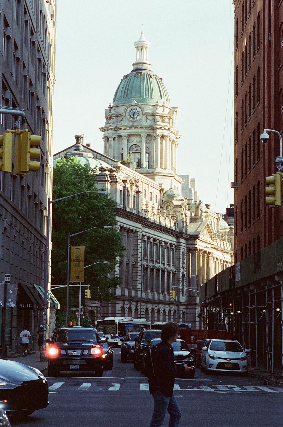 A large domed building with a clock tower stands prominently between two city buildings, with cars and a man on a street in the foreground.
