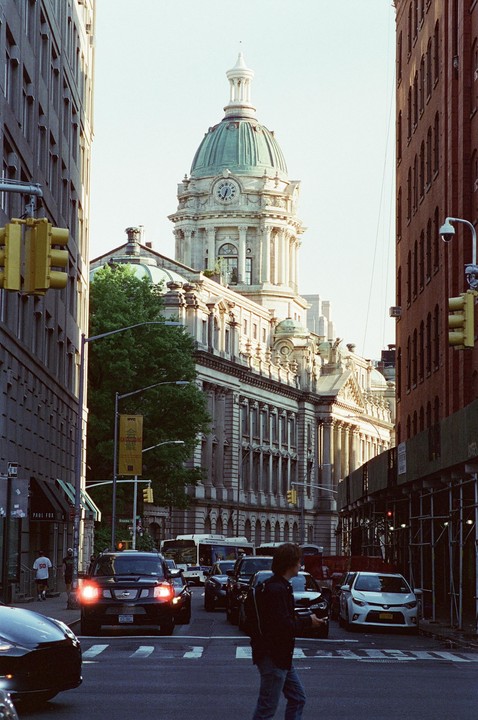 A large domed building with a clock tower stands prominently between two city buildings, with cars and a man on a street in the foreground.