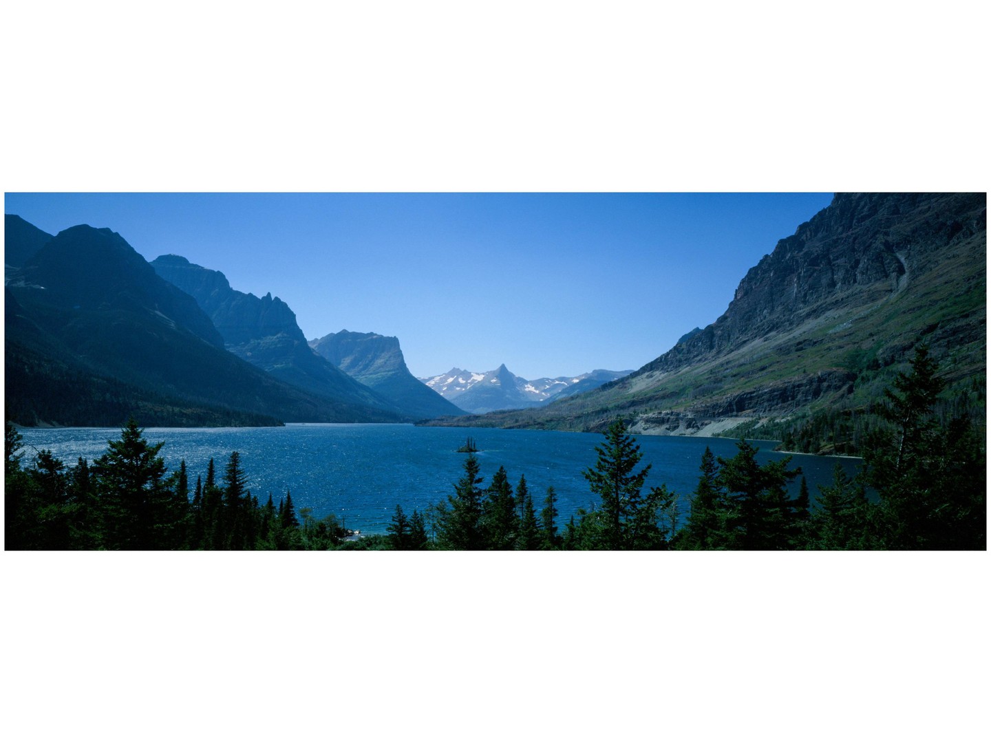 A wide shot of a lake surrounded by mountains and evergreen trees under a clear sky.