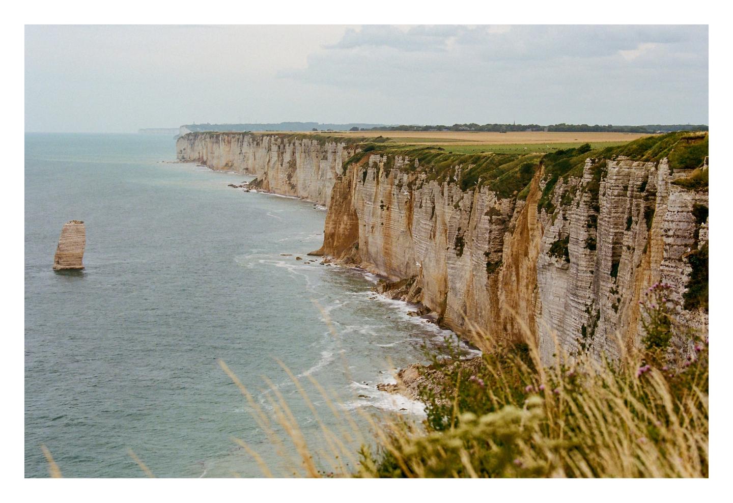 A long line of white cliffs with green tops stretches along the sea, with a single sea stack standing in the water.
