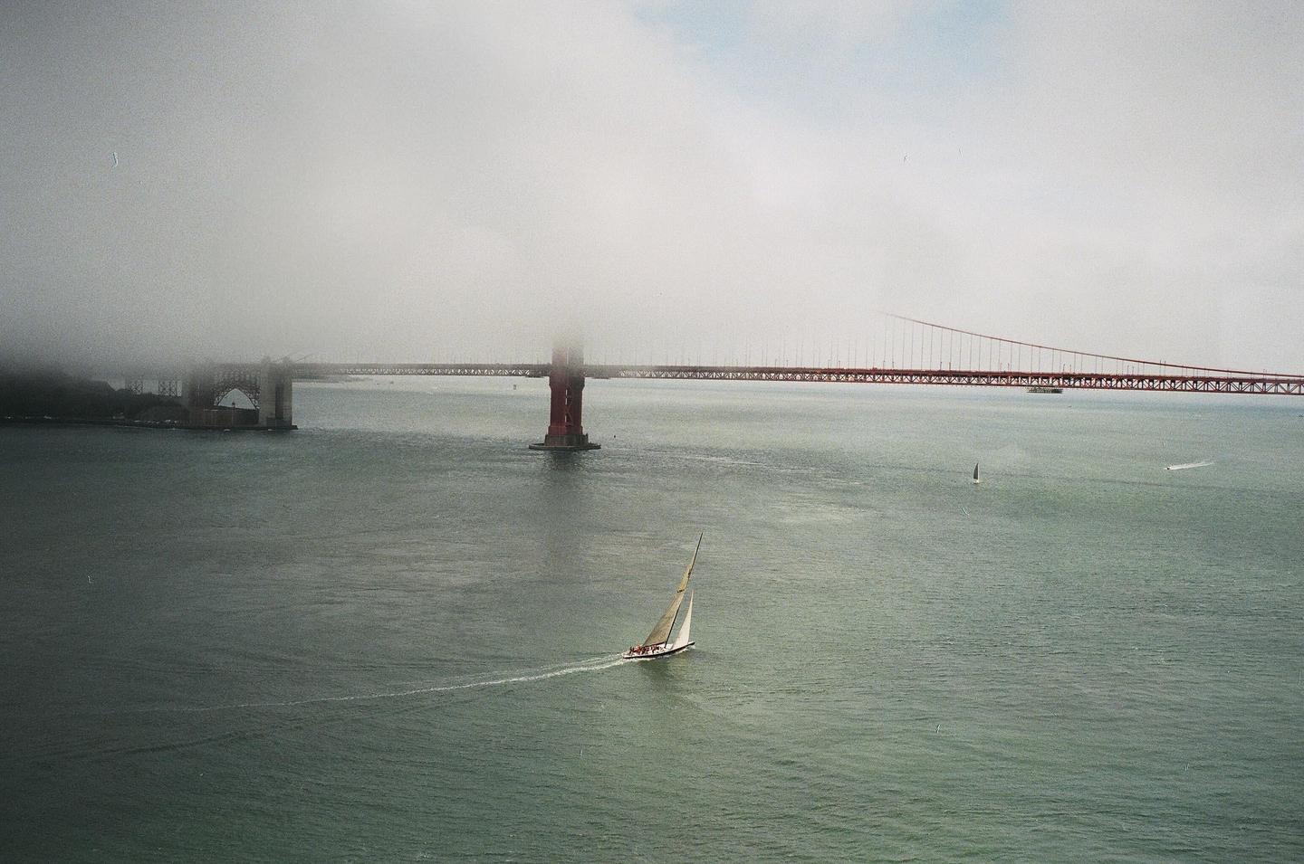 A sailboat moves across the water with the Golden Gate Bridge partially obscured by fog in the background.
