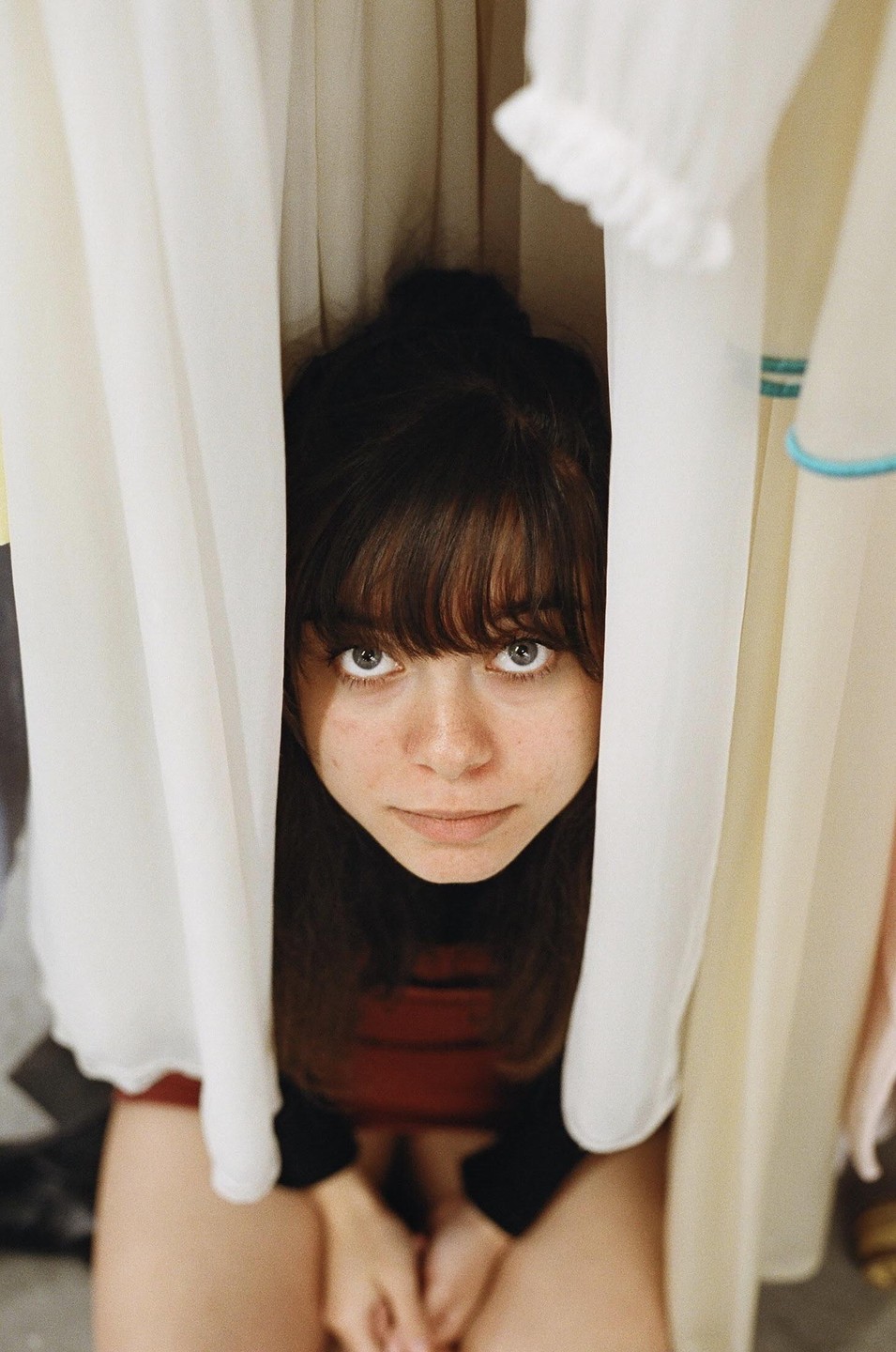 A young woman with bangs looks up at the camera from between two white curtains.