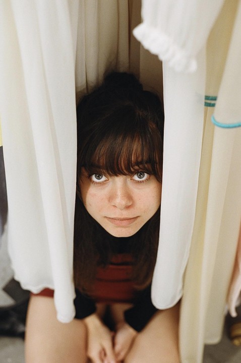 A young woman with bangs looks up at the camera from between two white curtains.