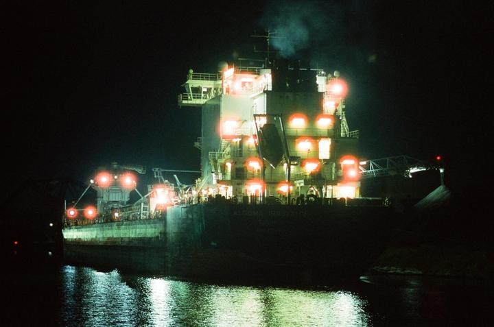 A large ship with many bright orange and white lights is docked at night, reflecting on the water.