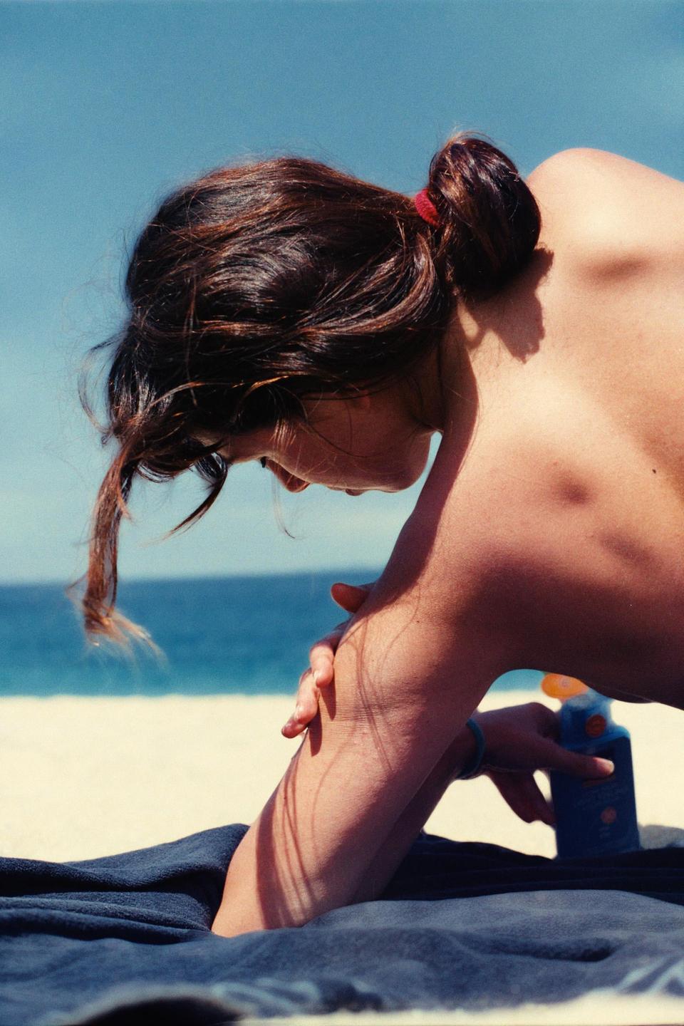 A woman applies sunscreen to her arm while lying on a towel on a beach.