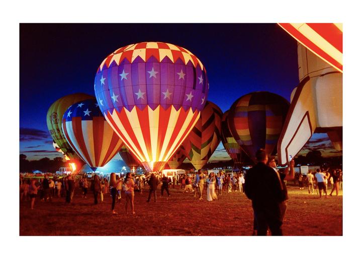 A crowd of people stands on a field at night, looking at several hot air balloons, one of which is shaped like a rocket.