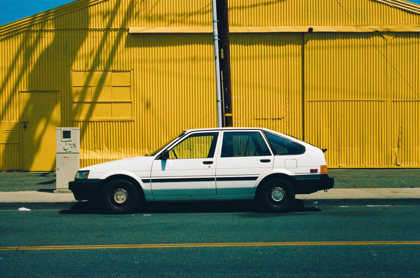 A white car is parked on a street next to a bright yellow corrugated building.