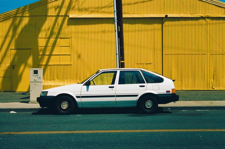 A white car is parked on a street next to a bright yellow corrugated building.