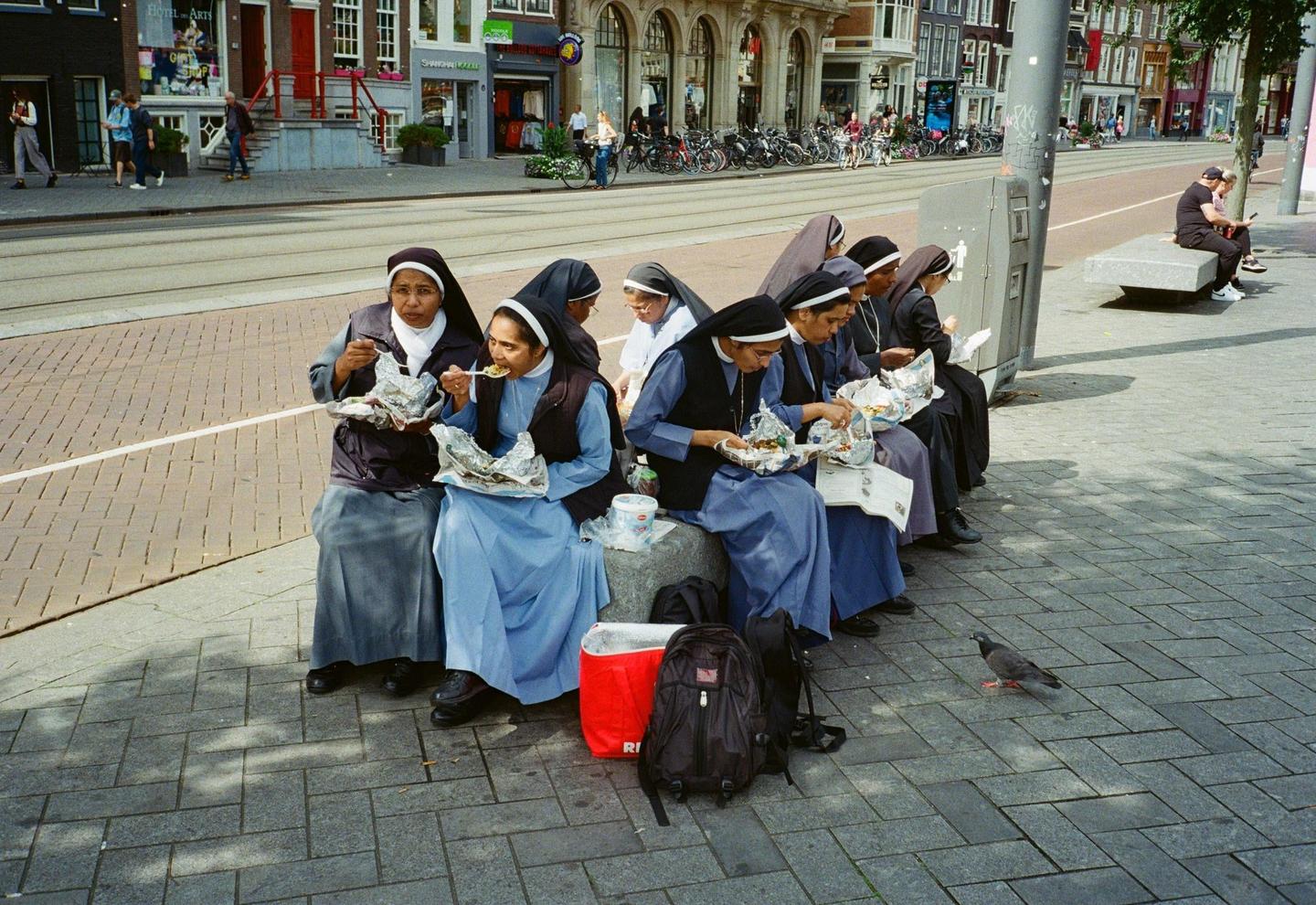 A group of nuns in blue and black habits are sitting on a stone bench on a city street, eating from foil-wrapped packages.