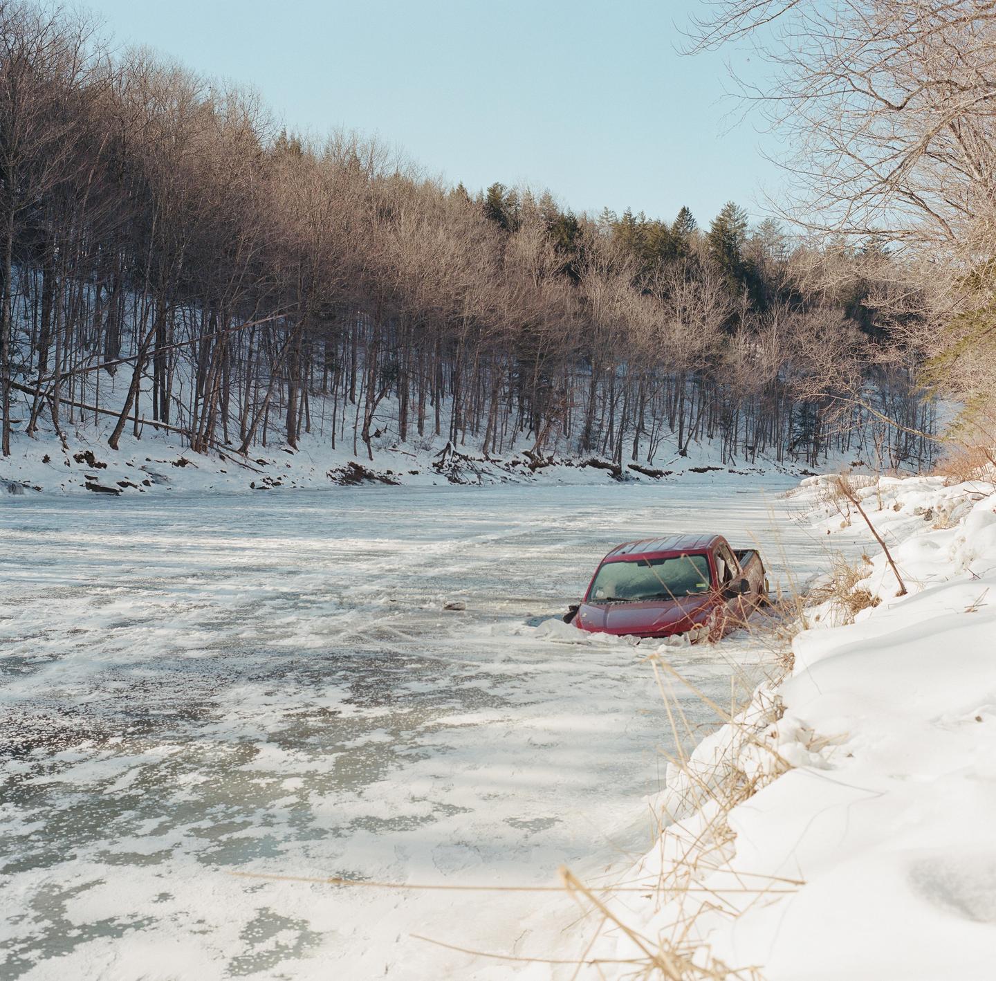 A red truck is partially submerged in a frozen river next to a snow-covered bank with a forest in the background.
