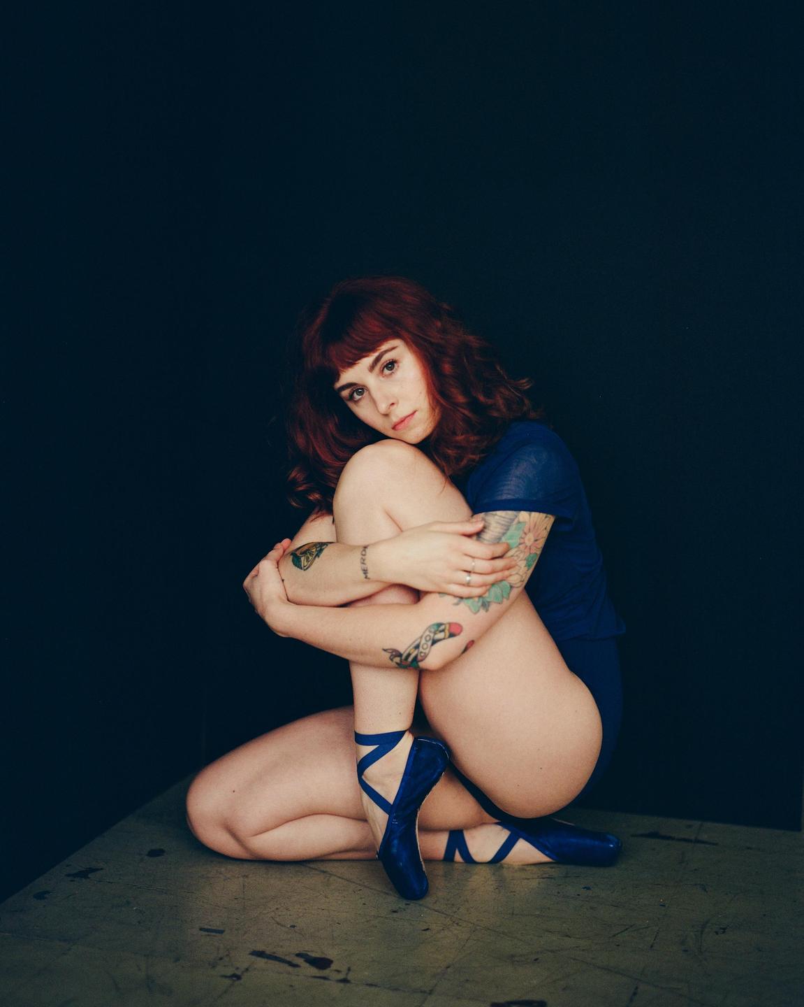 A woman with red hair and tattoos is sitting on a surface, wearing a blue leotard and blue ballet shoes.