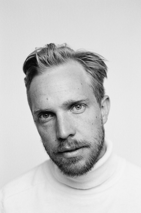 A man with a beard and a turtleneck sweater looks directly at the viewer.