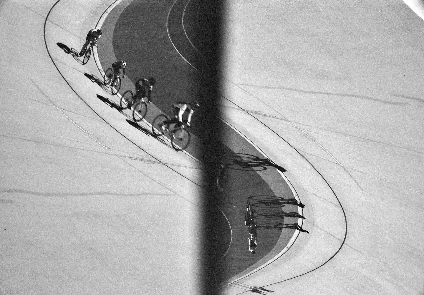 Four cyclists race on a velodrome track, casting long shadows.