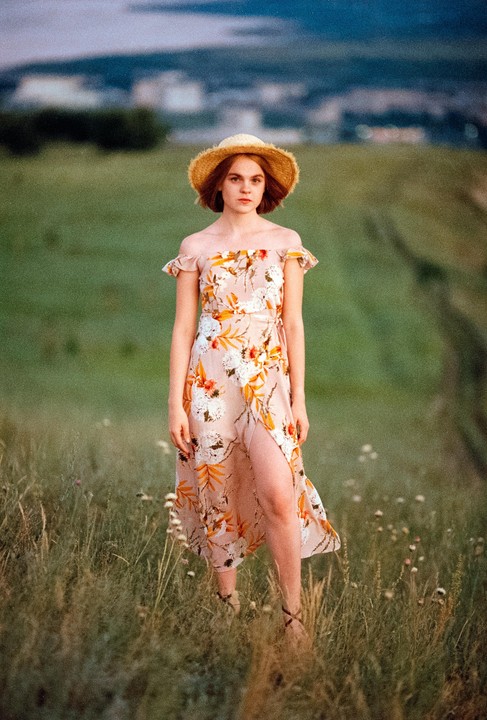 A woman in a floral dress and straw hat stands in a grassy field.