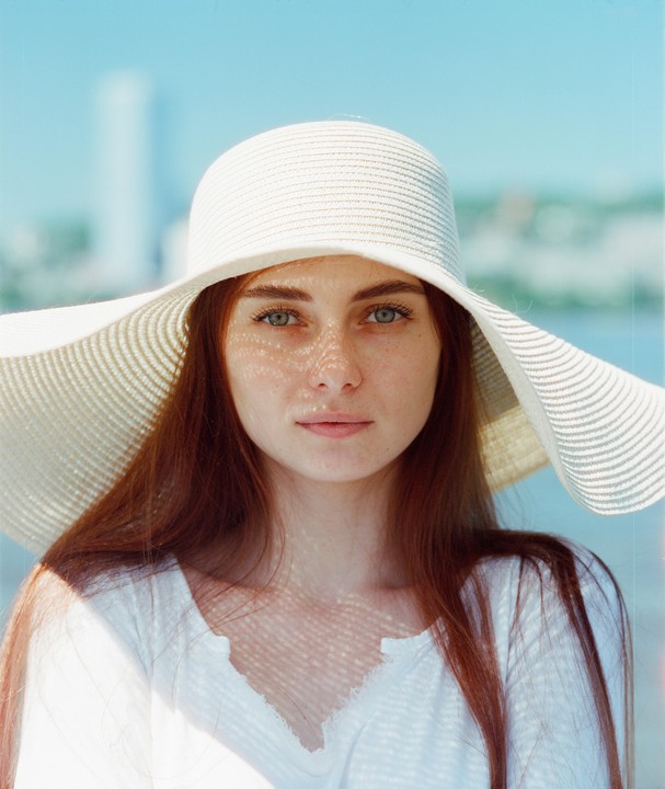 A woman with long red hair and a wide-brimmed hat looks directly at the viewer.