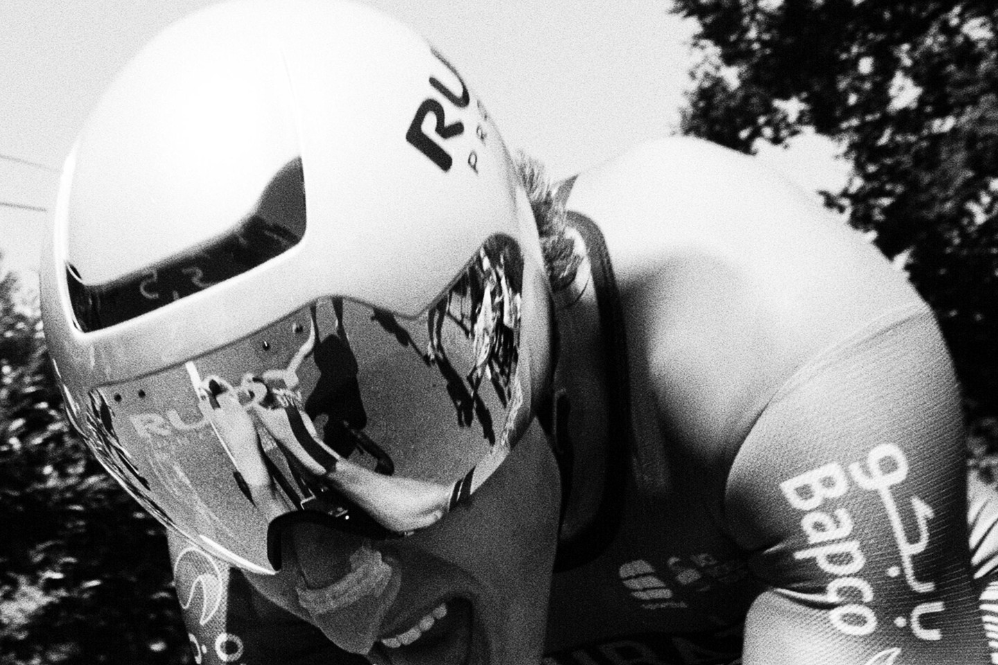 A cyclist wearing a helmet with a reflective visor is captured in a close-up, showing effort during a race.