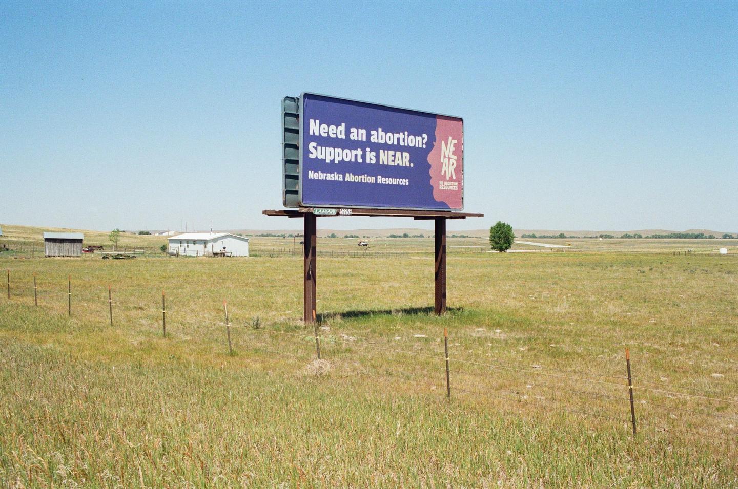 A billboard advertising Nebraska Abortion Resources stands in a grassy field under a clear sky.