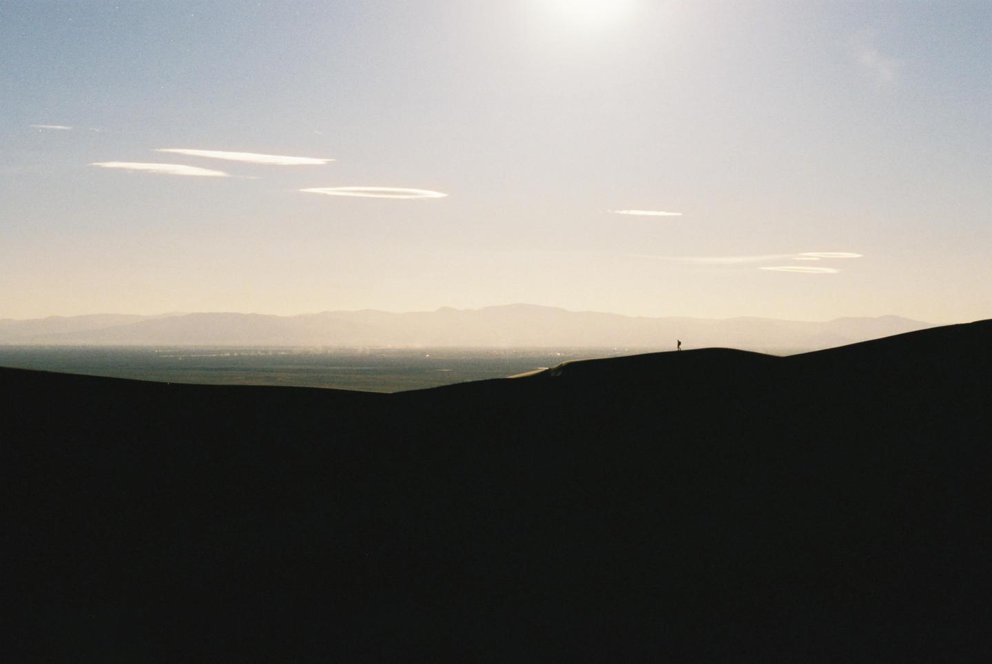 A person stands atop a sand dune with mountains and a valley in the distance under a bright sky.