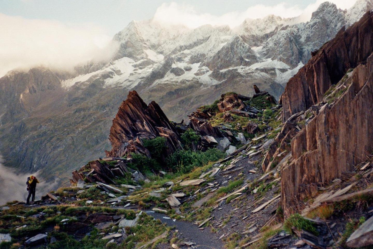 A person with a backpack stands on a rocky mountain path overlooking snow-capped peaks in the distance.