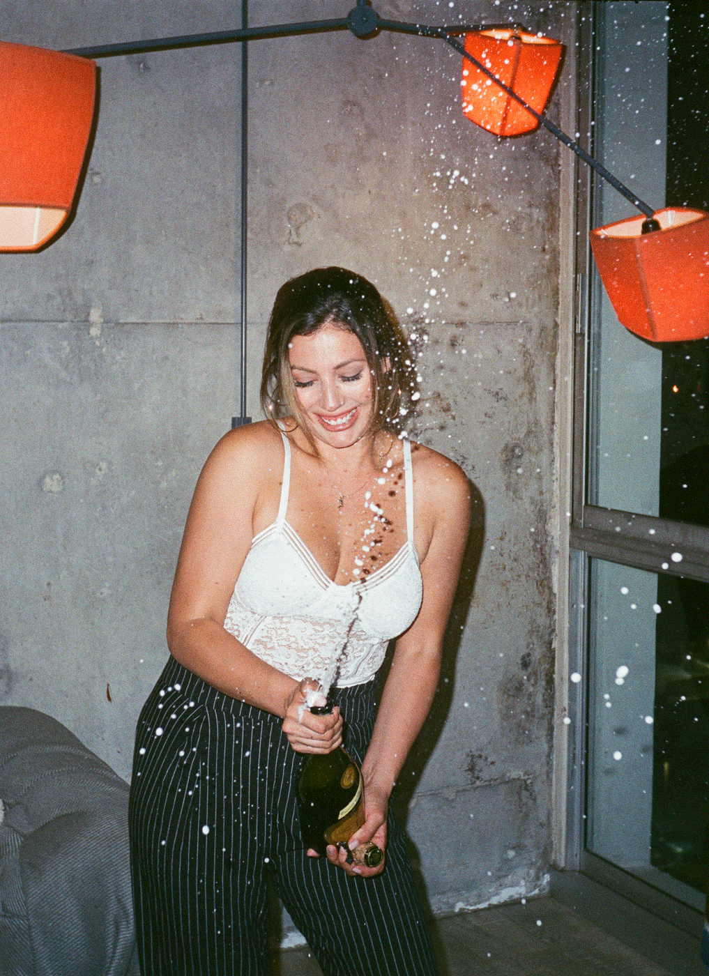 A woman in a white lace top and striped pants smiles as she opens a bottle of champagne, causing it to spray.