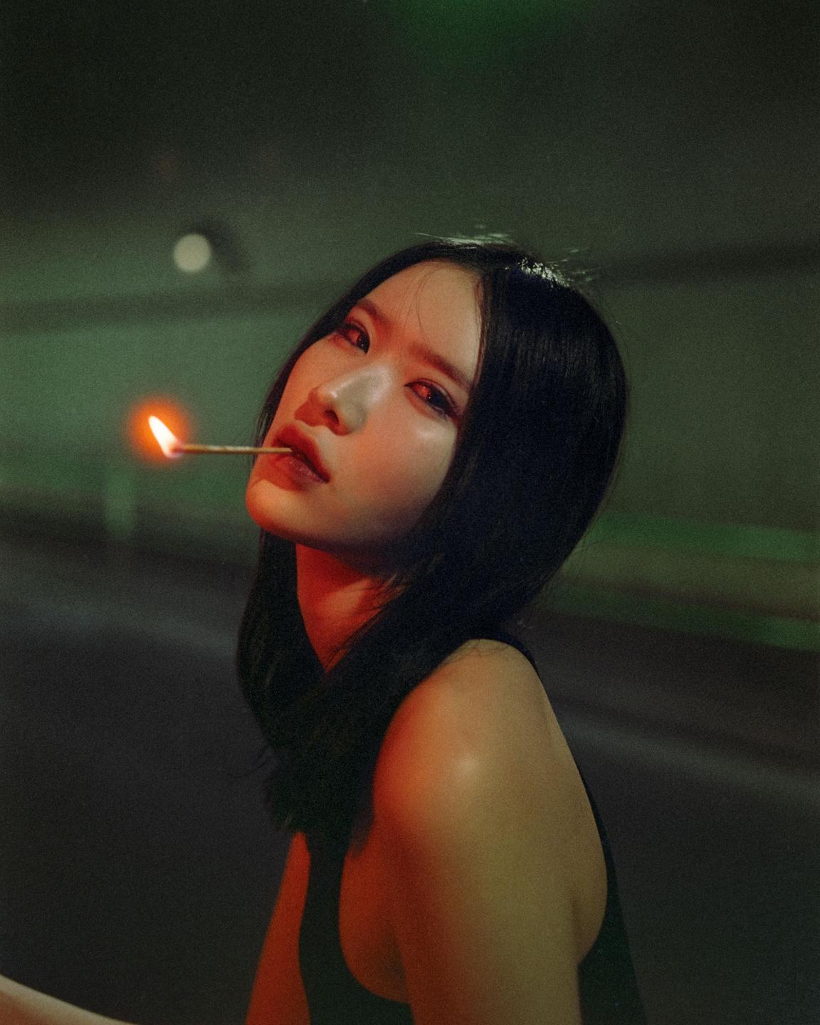 A woman holds a lit matchstick in her mouth in a dark setting.