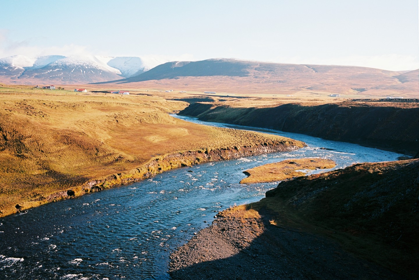A river winds through a vast, golden-brown landscape with snow-capped mountains in the distance under a clear sky.